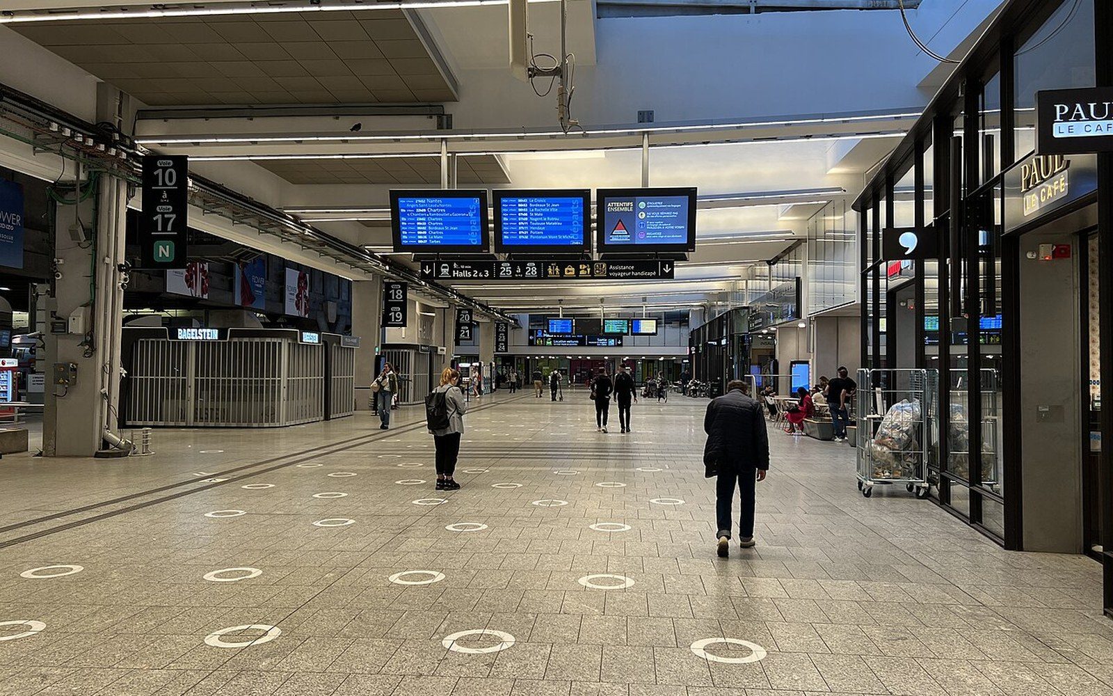 Travelers walk through a spacious train station concourse with departure boards and shops